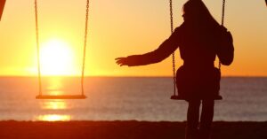 Woman on a swing at sunset reaching to an empty swing beside her